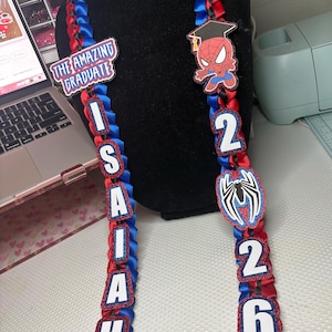 May include: Red and blue graduation leis with Spider-Man and graduation cap accents. One lei spells out the name "Isaiah" with white letters. The other lei has the numbers "2026". The text "The Amazing Graduate" is also visible.