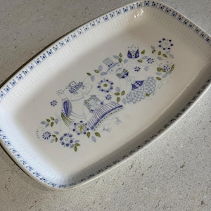 May include: Rectangular white ceramic serving platter with a blue floral and figure design. The platter has a blue patterned border and a central illustration of figures and flowers in blue, green, and purple.