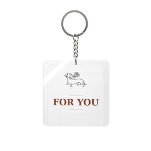 Peut inclure: Un porte-clés carré blanc avec une chaîne et un anneau argentés. Le porte-clés présente une illustration de teckel avec une rose et le texte "FOR YOU" en marron. Le design est simple et élégant.