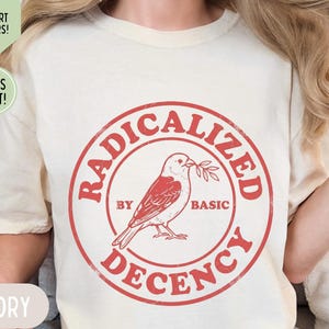 以下が含まれることがあります： アイボリー色のTシャツ。赤い円形のデザインで、「RADICALIZED BY BASIC DECENCY」の文字が小枝を持った鳥の周りに配置されています。 ゆったりとしたフィット感のシャツです。追加のテキストは、15色のシャツと迅速な発送を示しています。