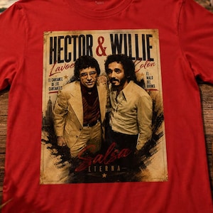 Può includere: Una t-shirt rossa a maniche corte con una stampa grafica di Hector Lavoe e Willie Colon. La stampa include il testo "HECTOR & WILLIE", "Lavoe", "Colon", "EL CANTANTE DE LOS CANTANTES", "EL MALO DEL BRONX" e "Salsa ETERNA" con un'icona a stella.