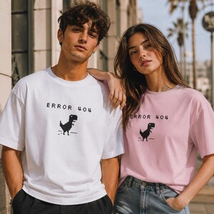 May include: White and pink t-shirts with the text "ERROR 404" and a pixelated dinosaur graphic. The t-shirts are short-sleeved and worn by two people. The background includes a building and palm trees.