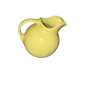 May include: A yellow ceramic pitcher with a rounded body and a curved handle. The pitcher has a spout for pouring and is set against a white background. The pitcher is a solid, pale yellow color.