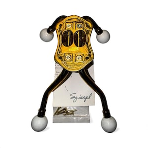 Pode incluir: Um cabide de parede de metal dourado com um design decorativo. O cabide tem dois braços com extremidades em forma de bola branca e um painel central com duas aberturas. Inclui uma etiqueta com o nome "Spiegel".