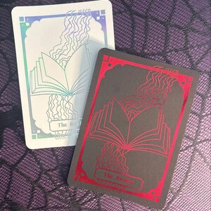 May include: Two tarot cards with a book and flowing lines design. One card has a white background with iridescent accents and the other has a black background with red accents. Both cards have the text "The Reader".