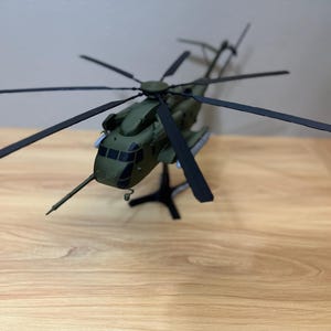 May include: A scale model of a military helicopter in olive green with black rotor blades. The aircraft features a prominent nose-mounted refueling probe, dark cockpit windows, and is mounted on a black tripod display stand for stability.