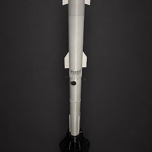 May include: A gray and white model rocket, standing upright on a black base. The rocket has a textured body, white fins, and a pointed nose cone. The base is a black, geometric shape.
