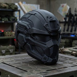 May include: A matte black tactical helmet featuring a full-face design with angular geometric paneling, a central forehead mounting bracket, integrated side rails, and a dark tinted visor that covers the eye area, providing a sleek, uniform appearance.