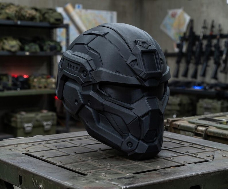 May include: A matte black tactical helmet featuring a full-face design with angular geometric paneling, a central forehead mounting bracket, integrated side rails, and a dark tinted visor that covers the eye area, providing a sleek, uniform appearance.