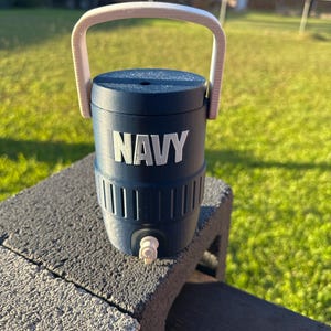 May include: A navy blue water cooler with a white handle and the word "NAVY" in white block letters. The cooler has a spigot and is sitting on a gray cinder block.