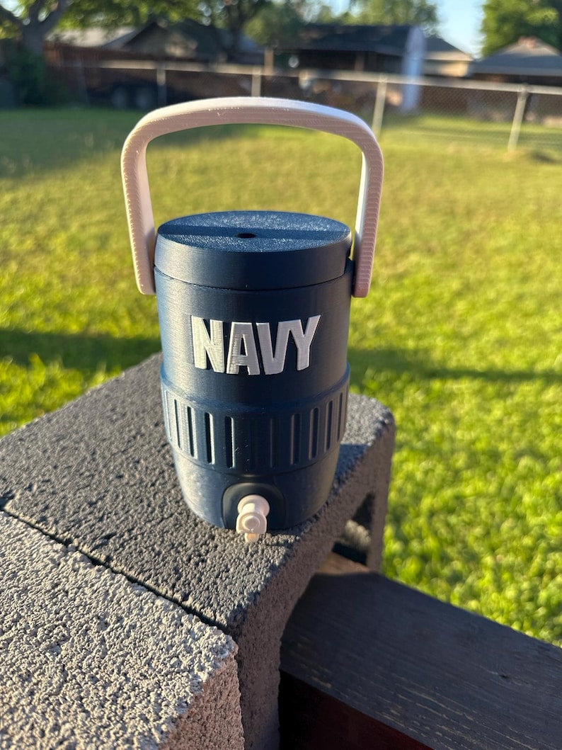 May include: A navy blue water cooler with a white handle and the word "NAVY" in white block letters. The cooler has a spigot and is sitting on a gray cinder block.