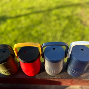 May include: Four colorful, cylindrical containers with handles. The containers are in olive green, red, gray, and navy blue. Each has a black top and a matching handle. The containers are sitting on a wooden surface with a green background.