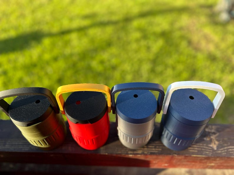 May include: Four colorful, cylindrical containers with handles. The containers are in olive green, red, gray, and navy blue. Each has a black top and a matching handle. The containers are sitting on a wooden surface with a green background.