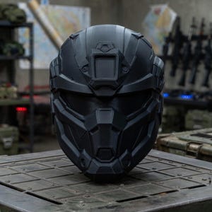 May include: A matte black tactical helmet featuring a geometric, multi-faceted design with a central forehead mount, a wide horizontal visor opening, and a reinforced chin guard. The helmet has a structured, angular shell with visible 3D print lines.