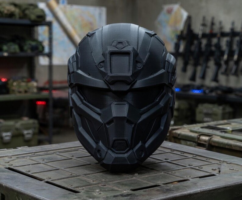 May include: A matte black tactical helmet featuring a geometric, multi-faceted design with a central forehead mount, a wide horizontal visor opening, and a reinforced chin guard. The helmet has a structured, angular shell with visible 3D print lines.