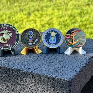 May include: Four commemorative coins on display, each mounted on a small stand. The coins represent the U.S. Marine Corps, U.S. Army, U.S. Air Force, and U.S. Navy. Each coin features detailed designs and text, including the 250th anniversary.
