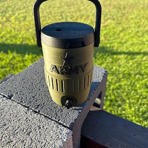 May include: Olive green water cooler with a black handle and lid. The word "ARMY" is printed in white, along with a star emblem. The cooler has a spigot and is sitting on a gray concrete block.
