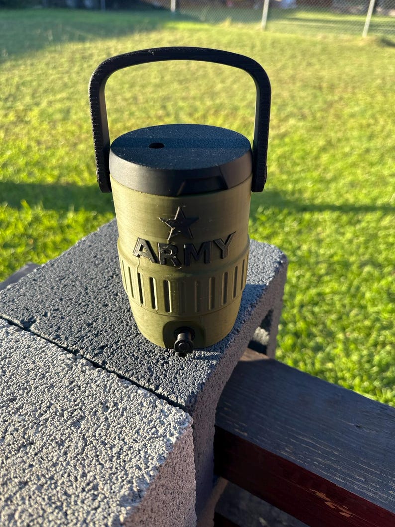 May include: Olive green water cooler with a black handle and lid. The word "ARMY" is printed in white, along with a star emblem. The cooler has a spigot and is sitting on a gray concrete block.
