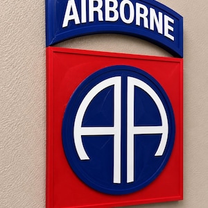 May include: A wall-mounted sign featuring the 82nd Airborne Division insignia. The design includes a red square base with a blue circle containing the white letters AA, topped by a blue arched tab with the white text AIRBORNE in bold letters.