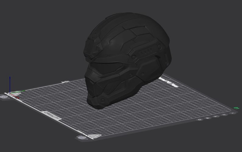 May include: A matte black tactical helmet with a geometric, multi-paneled design and a visor section, displayed as a 3D digital model on a grid-patterned build plate interface with visible software navigation icons and axis indicators.