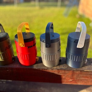 May include: Four small, cylindrical containers with handles, each in a different color: olive green, red, gray, and navy blue. The containers have a handle on top and vertical lines on the lower half. The olive green container has the word "ARK" printed on it.