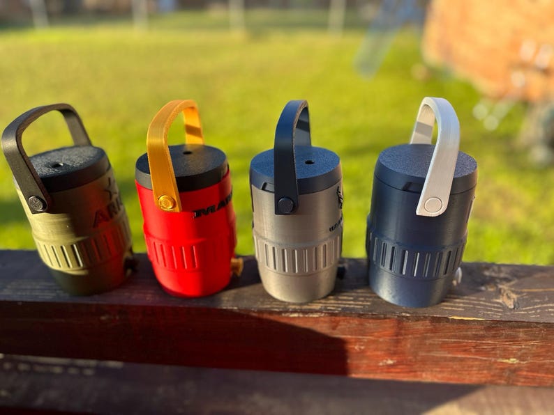 May include: Four small, cylindrical containers with handles, each in a different color: olive green, red, gray, and navy blue. The containers have a handle on top and vertical lines on the lower half. The olive green container has the word "ARK" printed on it.