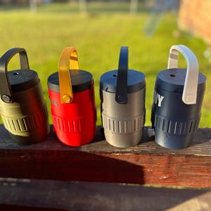 May include: Four cylindrical containers with handles in various colors: olive green, red, gray, and navy blue. Each container has a black top and a matching handle. The navy blue container has the word "NY" printed on it.