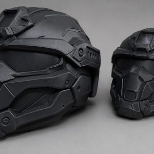 May include: Two matte black tactical helmets with a futuristic, angular design. Each helmet features a solid faceplate, a visor area, and a mounting bracket on the forehead. The helmets are constructed with layered, geometric panels and bolt details.