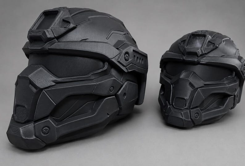 May include: Two matte black tactical helmets with a futuristic, angular design. Each helmet features a solid faceplate, a visor area, and a mounting bracket on the forehead. The helmets are constructed with layered, geometric panels and bolt details.