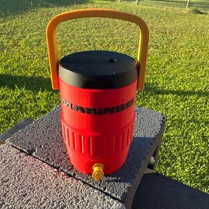 May include: A red water cooler with a black lid and a yellow handle. The word "MARINES" is printed on the side. It has a yellow spigot and is sitting on a gray surface.