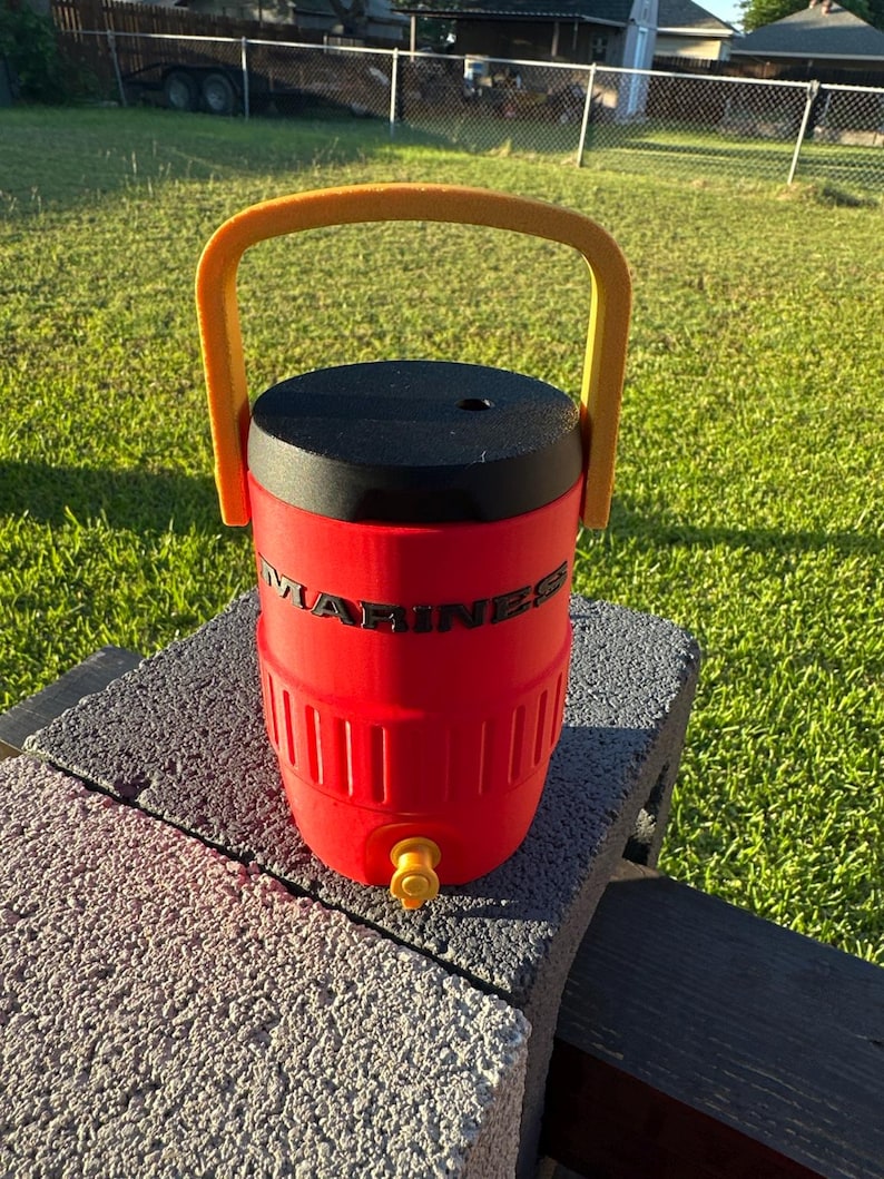 May include: A red water cooler with a black lid and a yellow handle. The word "MARINES" is printed on the side. It has a yellow spigot and is sitting on a gray surface.