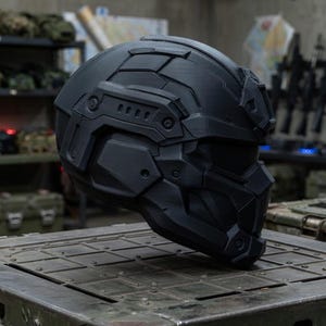 May include: A matte black tactical helmet featuring a full-face design with geometric paneling, integrated side rails, and a reinforced visor section. The helmet has a structured, multi-piece shell construction with visible screw details.