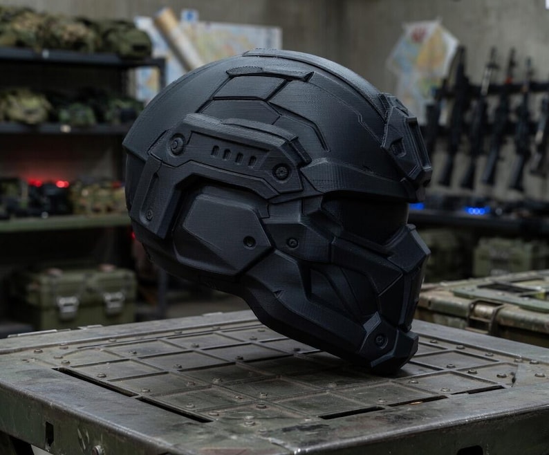 May include: A matte black tactical helmet featuring a full-face design with geometric paneling, integrated side rails, and a reinforced visor section. The helmet has a structured, multi-piece shell construction with visible screw details.