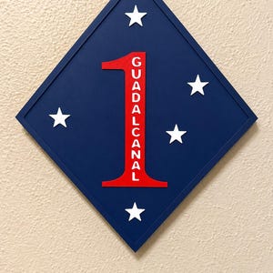 May include: A diamond-shaped blue wall plaque featuring a large red number one with the word GUADALCANAL printed vertically in white letters inside the number. The plaque is decorated with five white stars arranged around the central number.
