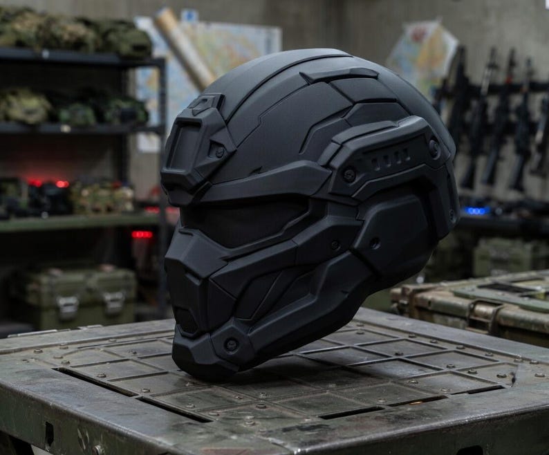 May include: A matte black tactical helmet featuring a full-face design with an integrated visor, a front-mounted accessory bracket, and segmented geometric paneling across the shell, providing a sleek, uniform appearance for protective gear.