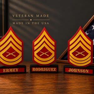 May include: Three red and yellow military rank insignias featuring crossed rifles, each mounted on a black base with a nameplate reading ERMEY, RODRIGUEZ, or JOHNSON. The text VETERAN MADE MADE IN THE USA is visible above the center insignia.