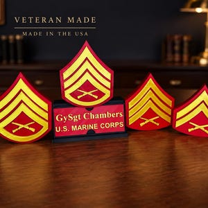 May include: A set of four red and gold U.S. Marine Corps Gunnery Sergeant rank insignias with crossed rifles. A black rectangular nameplate sits in the center with the text GySgt Chambers and U.S. MARINE CORPS in gold lettering.