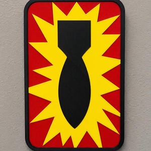 May include: A rectangular wall plaque featuring a black bomb silhouette centered over a bright yellow starburst shape, all set against a solid red background. The entire design is enclosed within a thin black frame with rounded corners.