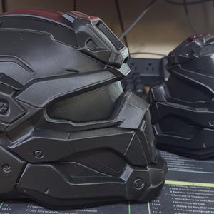 May include: Two matte black tactical helmets featuring a segmented, angular design with a prominent visor, raised ridges, and molded side vents. The helmets have a futuristic, armored appearance with detailed paneling and recessed screw-like accents.