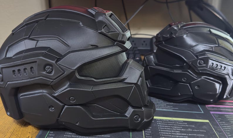 May include: Two matte black tactical helmets featuring a segmented, angular design with a prominent visor, raised ridges, and molded side vents. The helmets have a futuristic, armored appearance with detailed paneling and recessed screw-like accents.