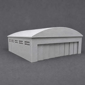 May include: A light gray scale model of a hangar building featuring a curved roof, a large front entrance with vertical panels, and a side wall with a row of four small rectangular windows. The structure has a textured surface finish throughout.