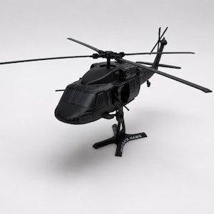 May include: A matte black scale model helicopter featuring a four-blade main rotor system and a tail rotor. The aircraft is mounted on a black cross-shaped display stand with the text BLACK HAWK printed in white on one of the base legs.