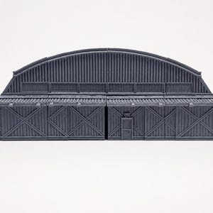 May include: A gray, 3D-printed miniature hangar building featuring a curved, corrugated roof and a modular facade with cross-braced wall panels. The structure includes a central rectangular door and horizontal window sections along the upper roofline.