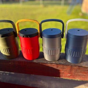 May include: Four colorful insulated tumblers with handles. The tumblers are olive green, red, gray, and navy blue. Each tumbler has a black or yellow handle and a matching lid. The tumblers are cylindrical with vertical grooves near the base.