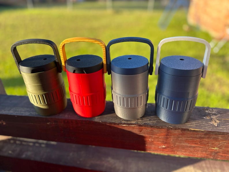 May include: Four colorful insulated tumblers with handles. The tumblers are olive green, red, gray, and navy blue. Each tumbler has a black or yellow handle and a matching lid. The tumblers are cylindrical with vertical grooves near the base.