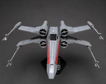 May include: A gray T-65B X-wing starfighter model with red accents on the fuselage, displayed on a black stand. The model features four engines and extended wings with laser cannons. The text "T-65B X-WING" is visible on the stand.