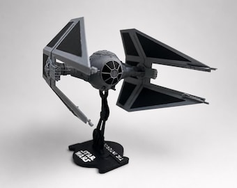 May include: A gray and black TIE Interceptor model starfighter from Star Wars, featuring a spherical cockpit with a black window pane design and four angled solar array wings. The model is mounted on a black display stand with Star Wars text.