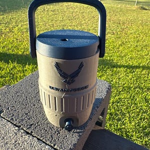 May include: A dark green water cooler with a dark blue lid and handle. The cooler features the US Air Force logo and text. It has a spigot at the bottom and is sitting on a gray brick.