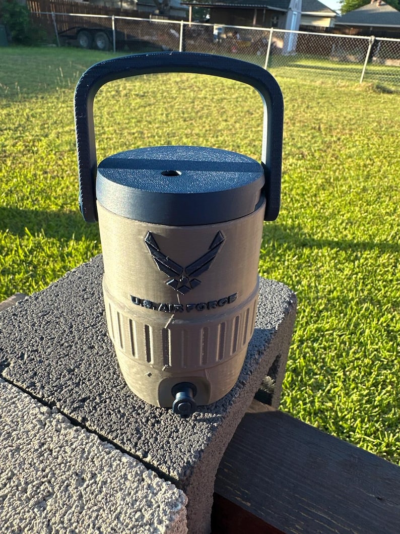 May include: A dark green water cooler with a dark blue lid and handle. The cooler features the US Air Force logo and text. It has a spigot at the bottom and is sitting on a gray brick.