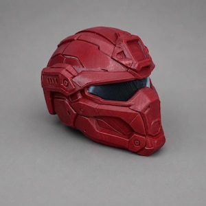 May include: A deep red tactical helmet featuring a segmented, multi-piece construction with a dark, recessed visor and a prominent rectangular mount on the forehead. The surface displays a textured finish with visible panel lines and geometric details.